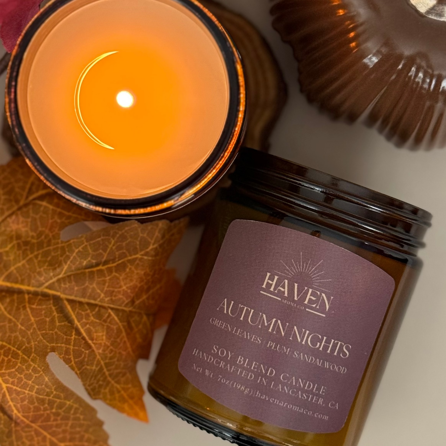Haven Autumn Nights candle with a lit candle and autumn leaves on a neutral background