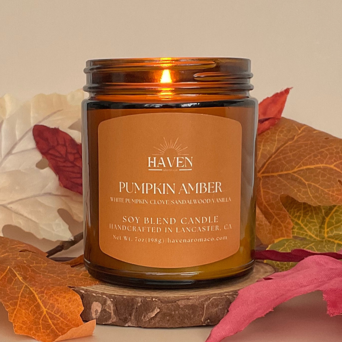 Haven Pumpkin Amber candle with a lit wick, surrounded by autumn leaves on a beige background.