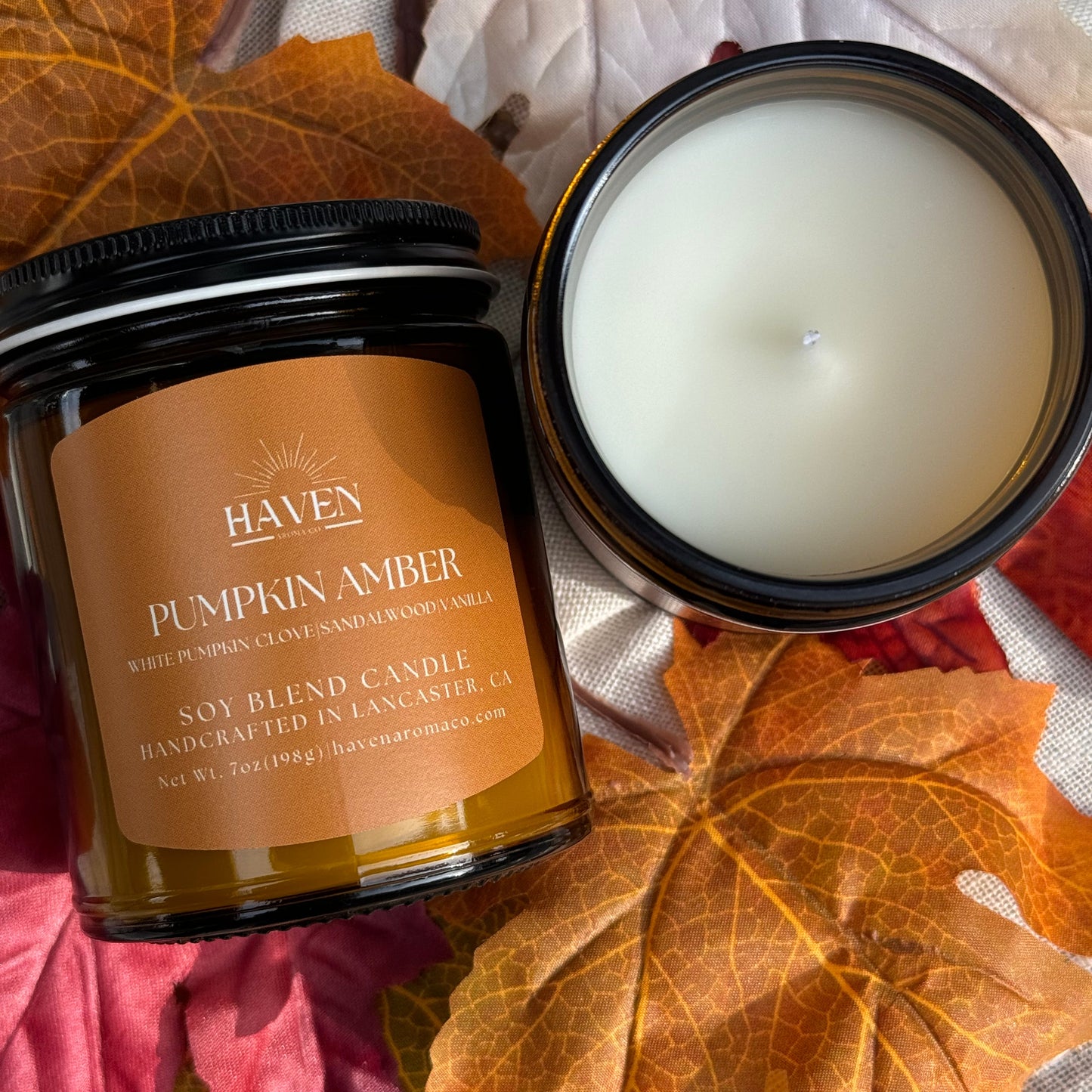 A 7oz pumpkin amber scented candle with its lid off, placed on a surface with autumn leaves surrounding it. The candle is labelled 'HAVEN AROMA CO.