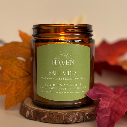 Candle labeled 'Fall Vibes' by Haven on a wooden coaster with autumn leaves in the background.
