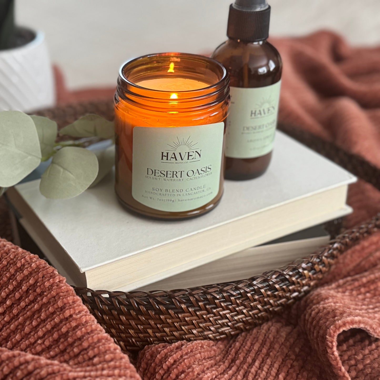 Haven Desert Oasis candle on a book with a bottle and eucalyptus leaves, surrounded by warm-toned fabric.
