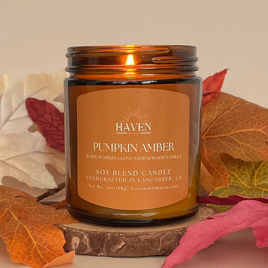 Haven Pumpkin Amber candle with a lit wick, surrounded by autumn leaves on a beige background.